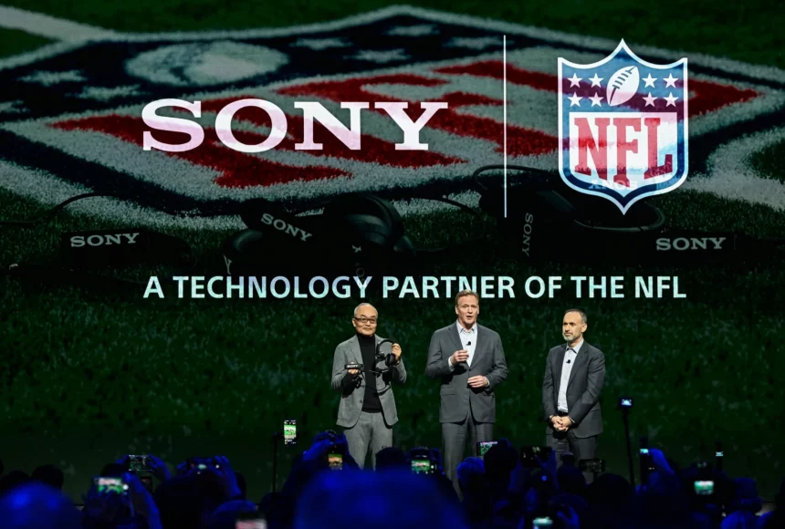 Drei Männer in Anzügen stehen auf einer Bühne vor großen SONY- und NFL-Logos, auf denen "A TECHNOLOGY PARTNER OF THE NFL" steht. Die Zuschauer halten Telefone für Fotos hoch, während Sony Headsets als wichtiges Hilfsmittel für NFL-Trainer in der Saison 2025 hervorgehoben werden. Diese Beschreibung wurde mit der FootballR KI automatisch generiert.