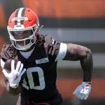 Quinshon Judkinsr in einer braunen Uniform und einem orangefarbenen Helm mit der Aufschrift "Judkins" läuft mit einem Football unter dem Arm während des Trainings. Quinshon Judkins' Handschuhe und sichtbare Tattoos stechen hervor, während er sich konzentriert auf das Spiel vorwärts bewegt. Diese Beschreibung wurde mit der FootballR KI automatisch generiert.