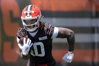 Quinshon Judkinsr in einer braunen Uniform und einem orangefarbenen Helm mit der Aufschrift "Judkins" läuft mit einem Football unter dem Arm während des Trainings. Quinshon Judkins' Handschuhe und sichtbare Tattoos stechen hervor, während er sich konzentriert auf das Spiel vorwärts bewegt. Diese Beschreibung wurde mit der FootballR KI automatisch generiert.