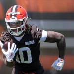 Quinshon Judkins in einer braunen Uniform und einem orangefarbenen Helm mit der Aufschrift "Judkins" läuft mit einem Football unter dem Arm während des Trainings. Quinshon Judkins' Handschuhe und sichtbare Tattoos stechen hervor, während er sich konzentriert auf das Spiel vorwärts bewegt. Diese Beschreibung wurde mit der FootballR KI automatisch generiert.