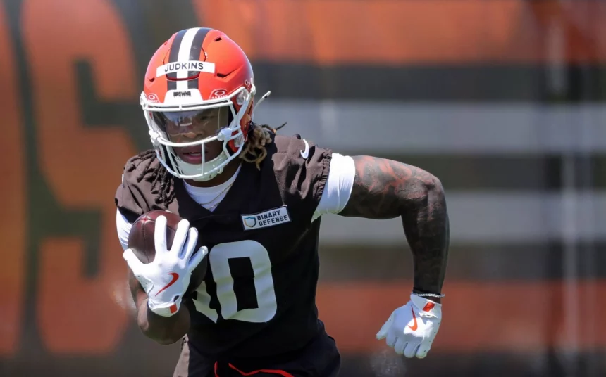 Quinshon Judkins in einer braunen Uniform und einem orangefarbenen Helm mit der Aufschrift "Judkins" läuft mit einem Football unter dem Arm während des Trainings. Quinshon Judkins' Handschuhe und sichtbare Tattoos stechen hervor, während er sich konzentriert auf das Spiel vorwärts bewegt. Diese Beschreibung wurde mit der FootballR KI automatisch generiert.