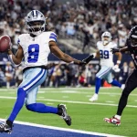 Der Spieler der Dallas Cowboys, KaVontae Turpin mit der Nummer 9, hält einen Football in der Hand und jubelt, als er in die Endzone läuft. Ein Falcons-Verteidiger in Schwarz und Rot versucht, ihn zu verfolgen, während im Hintergrund jubelnde Fans das Stadion füllen. Diese Beschreibung wurde mit der FootballR KI automatisch generiert.