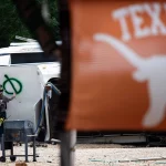 Ein Feuerwehrmann, der bei der Flutkatastrophe in Texas im Einsatz ist, sprüht einen grünen Kreis mit einer Linie auf ein umgestürztes Wohnmobil. Im Vordergrund prangt eine orangefarbene Texas Longhorns-Flagge mit der Aufschrift "TEXAS" inmitten von Bäumen und Sturmtrümmern. Diese Beschreibung wurde mit der FootballR KI automatisch generiert.