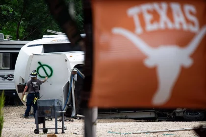 Ein Feuerwehrmann, der bei der Flutkatastrophe in Texas im Einsatz ist, sprüht einen grünen Kreis mit einer Linie auf ein umgestürztes Wohnmobil. Im Vordergrund prangt eine orangefarbene Texas Longhorns-Flagge mit der Aufschrift "TEXAS" inmitten von Bäumen und Sturmtrümmern. Diese Beschreibung wurde mit der FootballR KI automatisch generiert.
