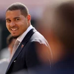 Ein Mann, Ryan Poles, trägt einen dunklen Anzug, ein helles Hemd und eine Krawatte und lächelt, während er über seine Schulter schaut. Er hat kurzes Haar und einen ordentlich gestutzten Bart. Der sanft verschwommene Hintergrund schafft eine professionelle und freundliche Atmosphäre. Diese Beschreibung wurde mit der FootballR KI automatisch generiert.