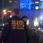 Eine Person, die eine schwarze Jacke mit dem gelben Schriftzug "FBI-NYPD JOINT TASK FORCE" trägt, steht nach einem Amoklauf auf einer nächtlichen Straße in New York. Unscharfe, blau beleuchtete Gebäude, Straßenlaternen und Barrikaden füllen den Hintergrund. Diese Beschreibung wurde mit der FootballR KI automatisch generiert.