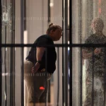 Eine Person mit Dienstmarke und zurückgekämmtem Haar beugt sich nach vorne und inspiziert das zerbrochene Glas eines nächtlichen Gebäudeeingangs, das möglicherweise mit einer Massenschießerei bei der NFL in Verbindung steht. Ihr Spiegelbild ist sichtbar; Barrikaden und ein Einschussloch markieren die angespannte Szene. Diese Beschreibung wurde mit der FootballR KI automatisch generiert.