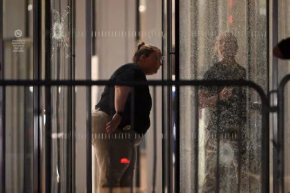 Eine Person mit Dienstmarke und zurückgekämmtem Haar beugt sich nach vorne und inspiziert das zerbrochene Glas eines nächtlichen Gebäudeeingangs, das möglicherweise mit einer Massenschießerei bei der NFL in Verbindung steht. Ihr Spiegelbild ist sichtbar; Barrikaden und ein Einschussloch markieren die angespannte Szene. Diese Beschreibung wurde mit der FootballR KI automatisch generiert.