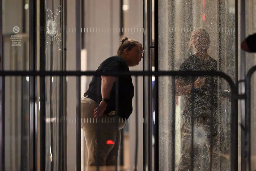 Eine Person mit Dienstmarke und zurückgekämmtem Haar beugt sich nach vorne und inspiziert das zerbrochene Glas eines nächtlichen Gebäudeeingangs, das möglicherweise mit einer Massenschießerei bei der NFL in Verbindung steht. Ihr Spiegelbild ist sichtbar; Barrikaden und ein Einschussloch markieren die angespannte Szene. Diese Beschreibung wurde mit der FootballR KI automatisch generiert.