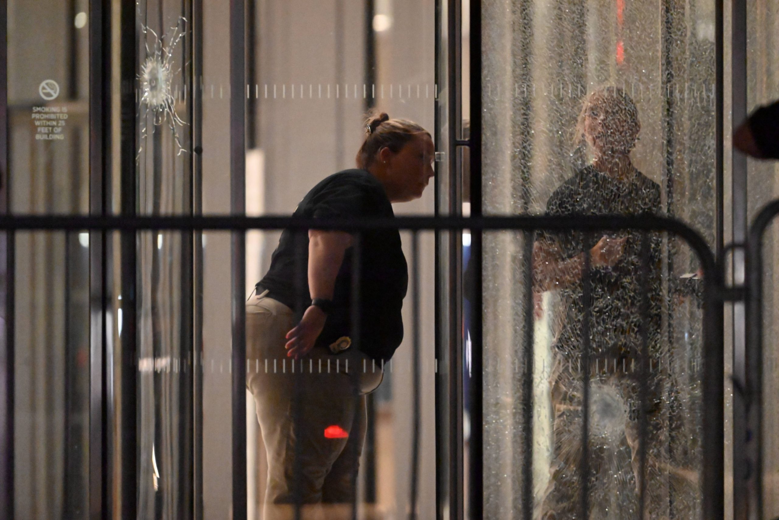 Eine Person mit Dienstmarke und zurückgekämmtem Haar beugt sich nach vorne und inspiziert das zerbrochene Glas eines nächtlichen Gebäudeeingangs, das möglicherweise mit einer Massenschießerei bei der NFL in Verbindung steht. Ihr Spiegelbild ist sichtbar; Barrikaden und ein Einschussloch markieren die angespannte Szene. Diese Beschreibung wurde mit der FootballR KI automatisch generiert.