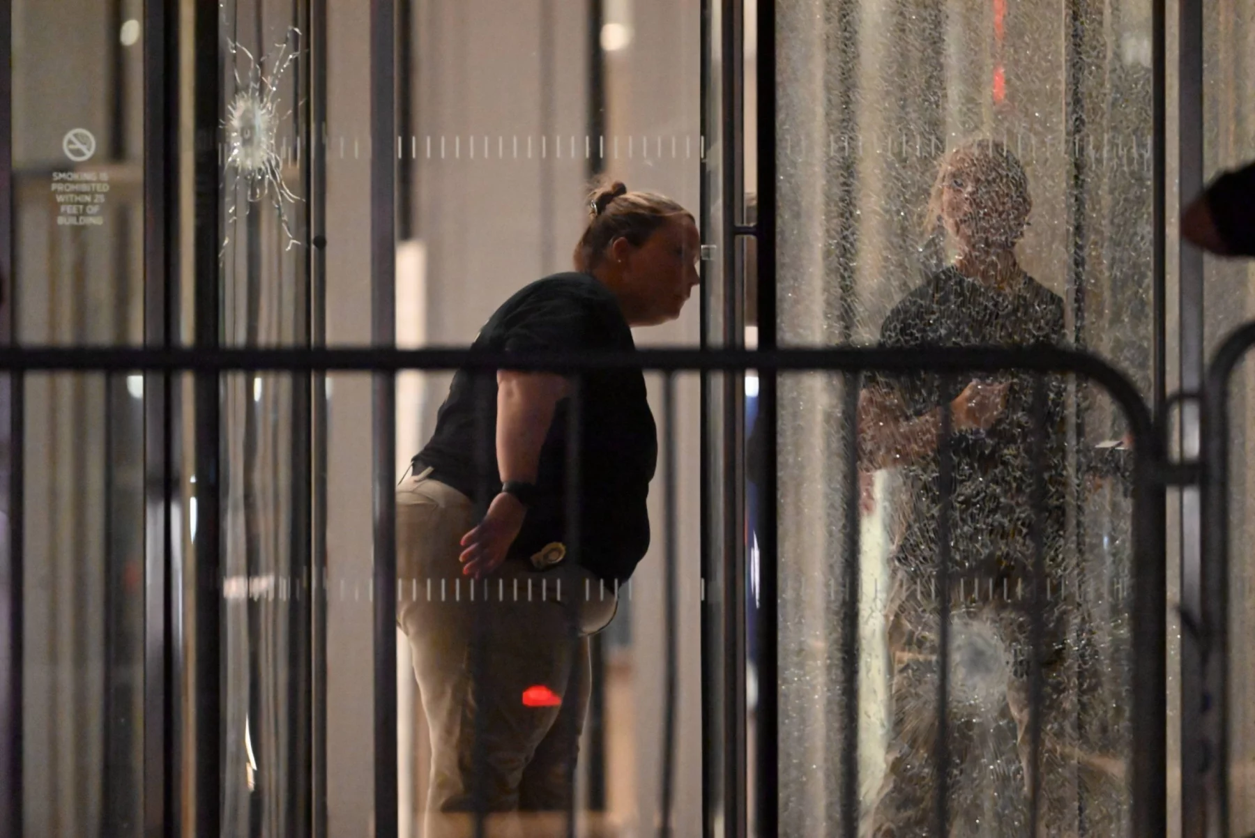 Eine Person mit Dienstmarke und zurückgekämmtem Haar beugt sich nach vorne und inspiziert das zerbrochene Glas eines nächtlichen Gebäudeeingangs, das möglicherweise mit einer Massenschießerei bei der NFL in Verbindung steht. Ihr Spiegelbild ist sichtbar; Barrikaden und ein Einschussloch markieren die angespannte Szene. Diese Beschreibung wurde mit der FootballR KI automatisch generiert.