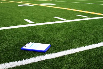 Ein blaues Klemmbrett mit Papieren liegt auf grünem Kunstrasen in der Nähe einer weißen Begrenzungslinie auf einem American-Football-Feld. Yard-Linien, darunter die Zahl 50, und gelbe Markierungen deuten auf ein NFL Dark Horse 2025 Training oder ein Spiel hin. Es sind keine Menschen anwesend. Diese Beschreibung wurde mit der FootballR KI automatisch generiert.