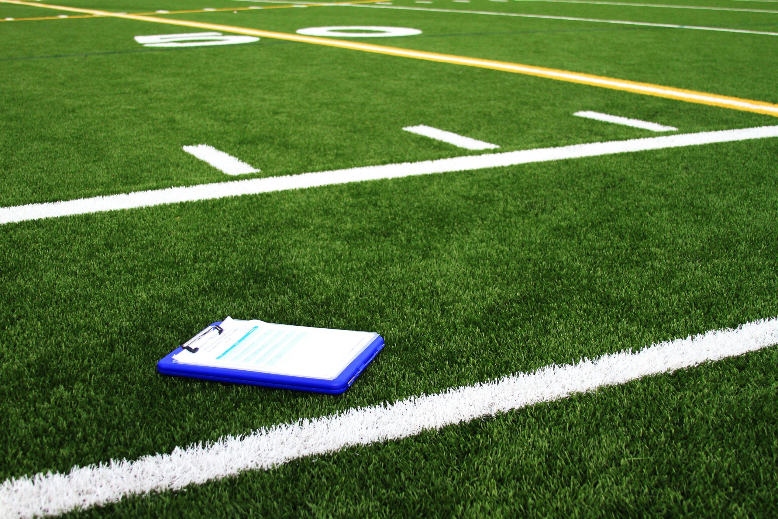 Ein blaues Klemmbrett mit Papieren liegt auf grünem Kunstrasen in der Nähe einer weißen Begrenzungslinie auf einem American-Football-Feld. Yard-Linien, darunter die Zahl 50, und gelbe Markierungen deuten auf ein NFL Dark Horse 2025 Training oder ein Spiel hin. Es sind keine Menschen anwesend. Diese Beschreibung wurde mit der FootballR KI automatisch generiert.
