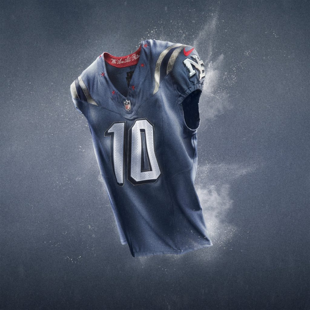Ein marineblaues NFL Rivalries-Trikots Football-Trikot mit silbernen und roten Akzenten schwebt vor einem grauen Hintergrund. Die Nummer 10 hebt sich in kräftigem Weiß ab, mit dem NFL-Logo unter dem Kragen und einem Patriots-Logo auf dem Ärmel inmitten eines dynamischen, rauchigen Effekts. Diese Beschreibung wurde mit der FootballR KI automatisch generiert.