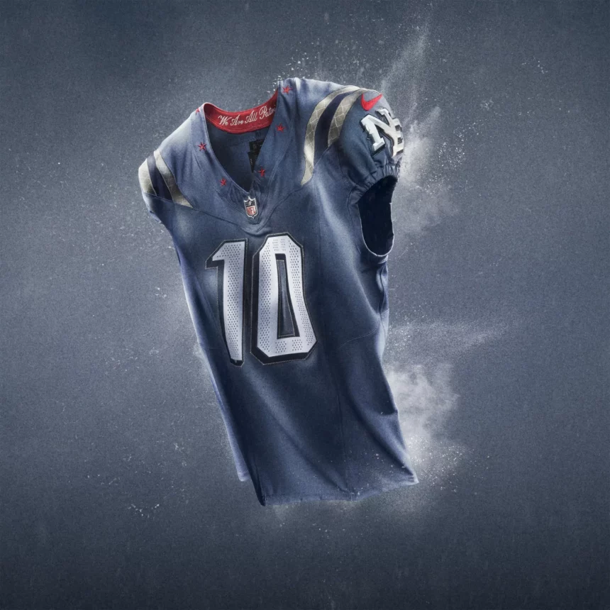 Ein marineblaues NFL Rivalries-Trikots Football-Trikot mit silbernen und roten Akzenten schwebt vor einem grauen Hintergrund. Die Nummer 10 hebt sich in kräftigem Weiß ab, mit dem NFL-Logo unter dem Kragen und einem Patriots-Logo auf dem Ärmel inmitten eines dynamischen, rauchigen Effekts. Diese Beschreibung wurde mit der FootballR KI automatisch generiert.