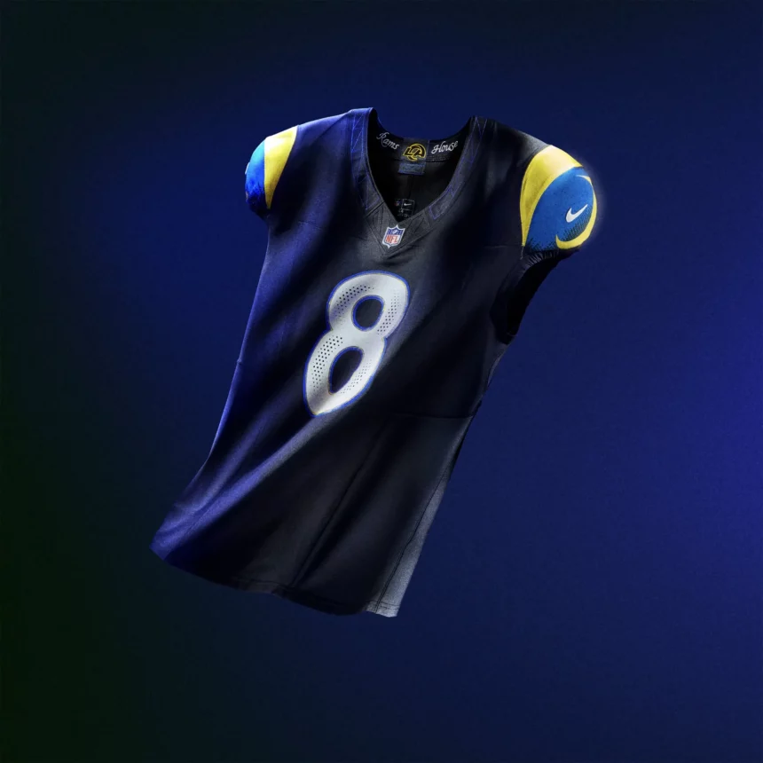 Ein Los Angeles Rams NFL Rivalries-Trikot mit der Nummer 8, dunkelblauen und leuchtend gelben Schulterakzenten sowie einem Nike-Logo auf dem Ärmel, das vor einem dunkelblauen und grünen Hintergrund mit Farbverlauf schwebt. Diese Beschreibung wurde mit der FootballR KI automatisch generiert.