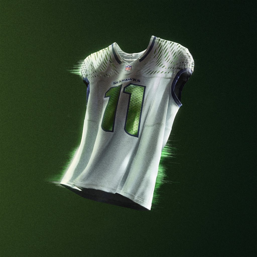 Ein weißes Seattle Seahawks NFL Rivalries-Trikot mit der Nummer 11 in Grün und Marineblau schwebt vor einem dunkelgrünen Hintergrund, wobei helle Lichter und Schatten seine Konturen und Details hervorheben. Diese Beschreibung wurde mit der FootballR KI automatisch generiert.