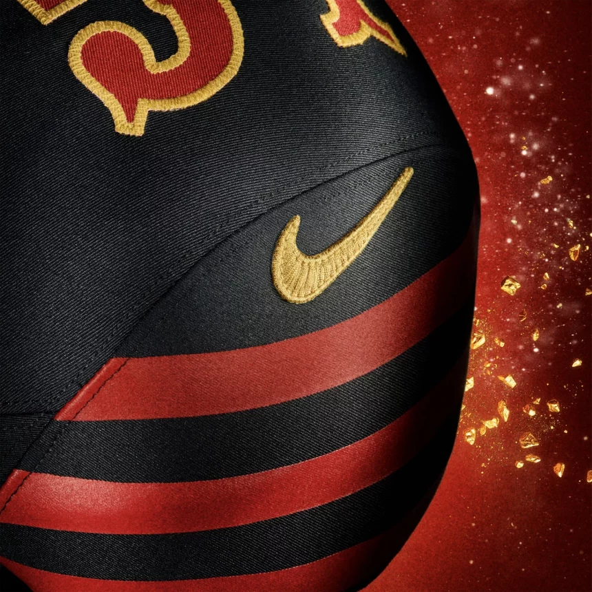 Eine Nahaufnahme eines schwarzen Sportballs mit roten und goldenen Details und einem goldenen Nike-Swoosh-Logo. Rote Streifen wickeln sich um den Ball, während goldenes Konfetti funkelt und die Energie der NFL Rivalries-Trikots vor einem leuchtend roten Hintergrund einfängt. Diese Beschreibung wurde mit der FootballR KI automatisch generiert.