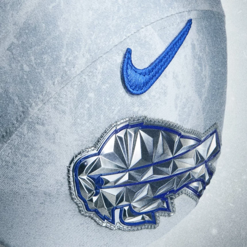 Die Nahaufnahme einer hellgrauen Baseballkappe zeigt ein blaues Nike-Swoosh-Logo über einem glänzenden, silbernen, geometrischen Büffelaufnäher, der blau umrandet ist und einen Hauch von NFL Rivalries-Trikots vermittelt. Die Kappe hat eine weiche, leicht verblasste Stofftextur. Diese Beschreibung wurde mit der FootballR KI automatisch generiert.
