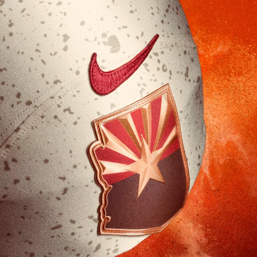 Ein weiß gesprenkelter Sporthelm mit einem roten Nike-Swoosh und einem Aufnäher in Form von Arizona, der einen stilisierten Starburst in den Farben Rot, Gold und Braun zeigt - perfekt, um die NFL-Rivalitäten zu feiern - Trikots. Der Hintergrund ist ein strukturiertes, leuchtendes Orange. Diese Beschreibung wurde mit der FootballR KI automatisch generiert.