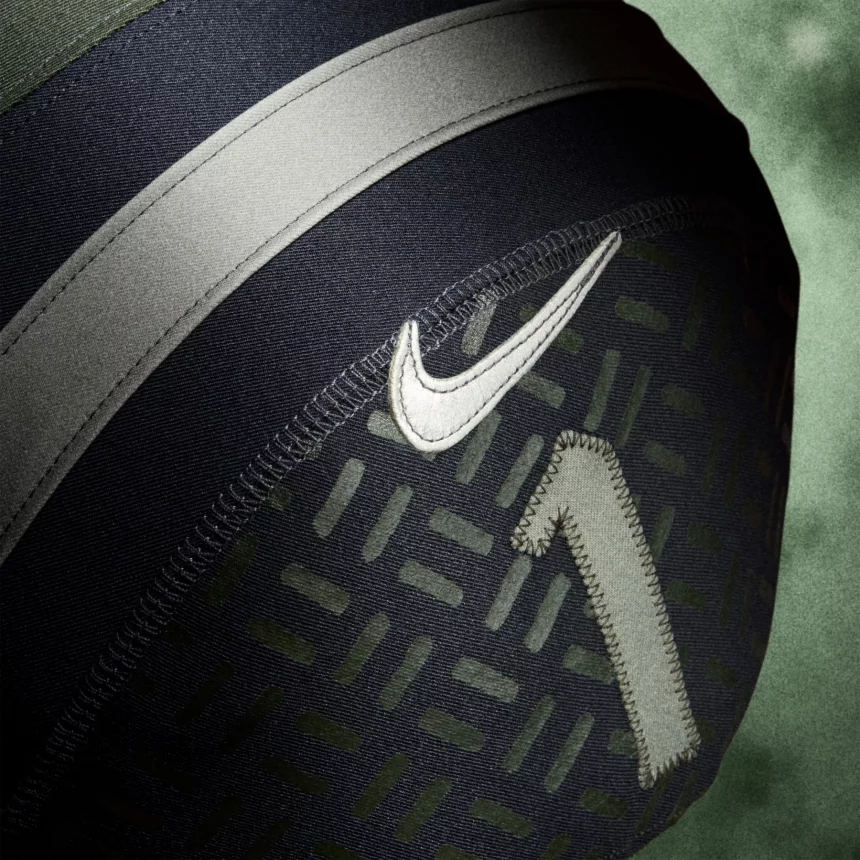 Nahaufnahme eines dunkelgrünen und schwarzen Footballs mit einem aufgestickten weißen Nike-Swoosh-Logo und einer großen aufgestickten Nummer 1. Die gemusterte Textur des Balls und der weiße Streifen erinnern an die Intensität der NFL Rivalries-Trikots vor einem grünen, verschwommenen Hintergrund. Diese Beschreibung wurde mit der FootballR KI automatisch generiert.