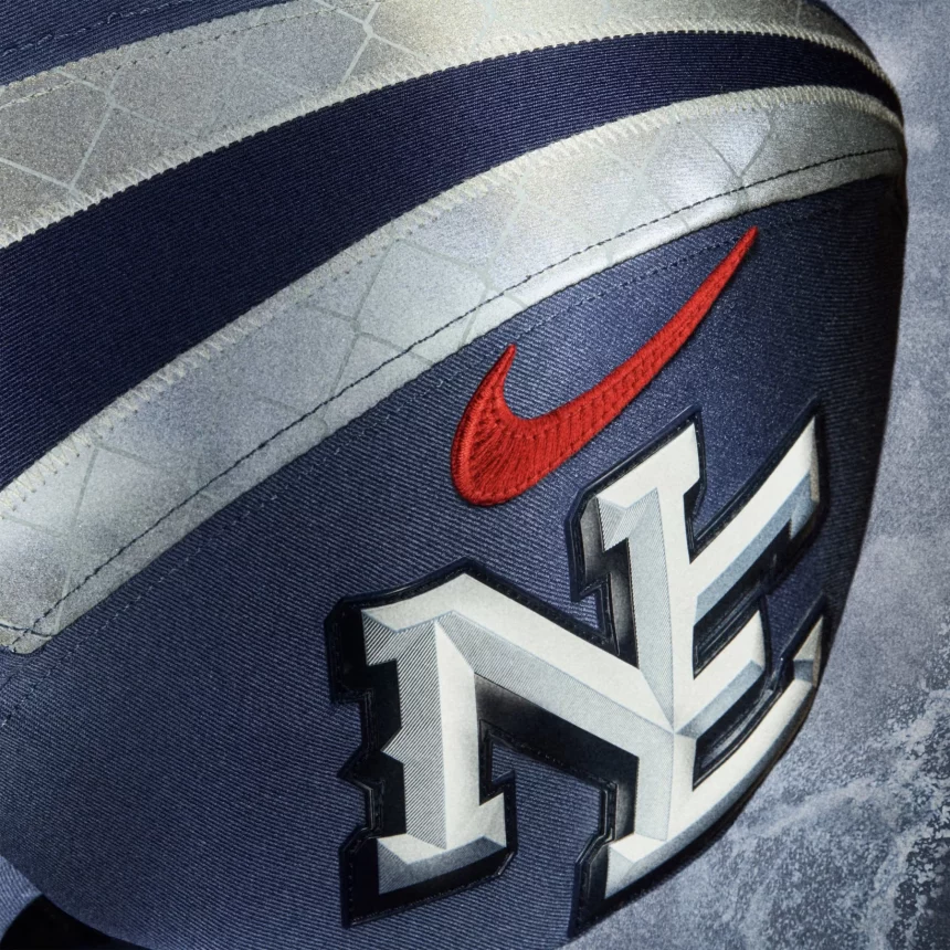 Nahaufnahme eines navyblauen Footballs mit rotem Nike-Swoosh und auffälligem "NE"-Logo, das an NFL-Rivalries-Trikots erinnert. Die strukturierte Oberfläche, die detaillierten Nähte und der silberne Streifen, der sich über die Oberseite schlängelt, unterstreichen das hochwertige Design. Diese Beschreibung wurde mit der FootballR KI automatisch generiert.