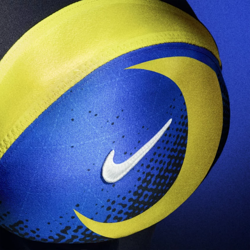 Nahaufnahme eines blauen und gelben Nike-Fußballs mit einem weißen Swoosh-Logo. Das kühne Design, das an die NFL Rivalries-Trikots erinnert, zeigt gelbe Kurven und gepunktete Akzente auf blauem Grund, wobei die Oberflächenstruktur des Balls deutlich sichtbar ist. Diese Beschreibung wurde mit der FootballR KI automatisch generiert.
