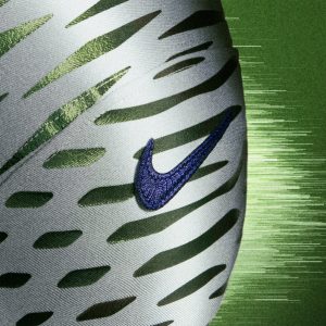 Nahaufnahme eines weißen Sportschuhs mit grünen Schlitzen, einem dunkelblauen Nike-Swoosh-Logo und einem strukturierten grünen Hintergrund, der an NFL Rivalries-Trikots erinnert, mit verschwommenen horizontalen Streifen auf der rechten Seite, die ein Gefühl der Bewegung vermitteln. Diese Beschreibung wurde mit der FootballR KI automatisch generiert.