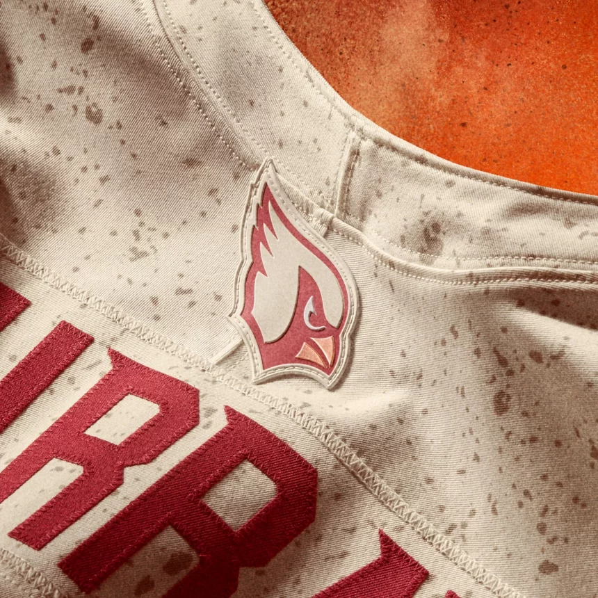 Nahaufnahme eines cremefarbenen NFL Rivalries-Trikots mit roten Sprenkeln, auf dem das Logo der Arizona Cardinals in großen, fetten roten Buchstaben zu sehen ist. Im Hintergrund erscheint ein Teil eines orangefarbenen Football-Feldes oder -Platzes. Diese Beschreibung wurde mit der FootballR KI automatisch generiert.