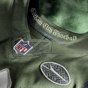 Nahaufnahme eines grünen NFL Rivalries-Trikots mit aufgenähtem "Gotham City Football" am Kragen, einem NFL-Logoaufnäher und einem kreisförmigen Fledermaus-Silhouetten-Emblem auf gemustertem Hintergrund vor einem grünen, verschwommenen Hintergrund. Diese Beschreibung wurde mit der FootballR KI automatisch generiert.