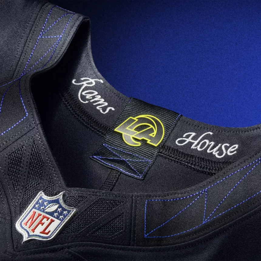Nahaufnahme des Kragens eines Los Angeles Rams NFL Rivalries-Trikots. Der Kragen zeigt den Schriftzug "Rams House" in weißer Schrift, ein Rams-Logo-Tag, blaue Nähte und einen NFL-Schildaufnäher auf schwarzem Stoff, alles vor blauem Hintergrund. Diese Beschreibung wurde mit der FootballR KI automatisch generiert.
