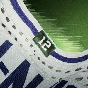 Nahaufnahme eines NFL Rivalries-Trikots mit der Nummer 12 auf einem grünen rechteckigen Aufnäher am Halsausschnitt. Das weiße Trikot hat blaue Details, Belüftungslöcher entlang des Kragens und einen grünen Hintergrund mit Farbverlauf und leichten Streifeneffekten. Diese Beschreibung wurde mit der FootballR KI automatisch generiert.