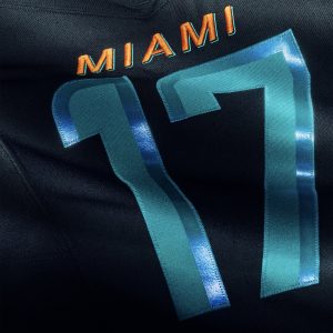 Nahaufnahme eines dunklen NFL Rivalries-Trikots mit dem orangefarbenen Schriftzug "MIAMI" über der großen, glänzenden blaugrünen Nummer 17. Die Textur des Stoffes und die Details der Nähte sind sichtbar, wobei das Licht von den glatten Zahlen reflektiert wird. Diese Beschreibung wurde mit der FootballR KI automatisch generiert.