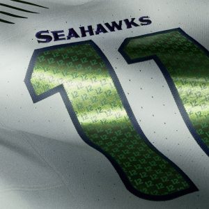 Nahaufnahme eines weißen NFL Rivalries-Trikots mit der Aufschrift "SEAHAWKS" in fetten blauen und grünen Buchstaben über einer leuchtend grünen Nummer 12. Das Muster aus winzigen "12 "s im Inneren unterstreicht die leidenschaftliche Fangemeinde der Seattle Seahawks. Diese Beschreibung wurde mit der FootballR KI automatisch generiert.