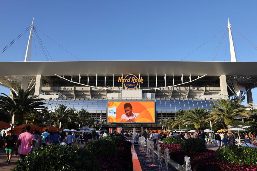 Miami Dolphins - Eine Menschenmenge versammelt sich an einem sonnigen Tag vor dem Hard Rock Stadium. Palmen, Springbrunnen und eine große Leinwand, auf der Ryan Crow in einem weißen Hemd zu sehen ist, ragen unter dem ikonischen Dach des Stadions und dem "Hard Rock Stadium"-Schild in den klaren blauen Himmel. Diese Beschreibung wurde mit der FootballR KI automatisch generiert.