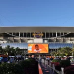 Miami Dolphins - Eine Menschenmenge versammelt sich an einem sonnigen Tag vor dem Hard Rock Stadium. Palmen, Springbrunnen und eine große Leinwand, auf der Ryan Crow in einem weißen Hemd zu sehen ist, ragen unter dem ikonischen Dach des Stadions und dem "Hard Rock Stadium"-Schild in den klaren blauen Himmel. Diese Beschreibung wurde mit der FootballR KI automatisch generiert.