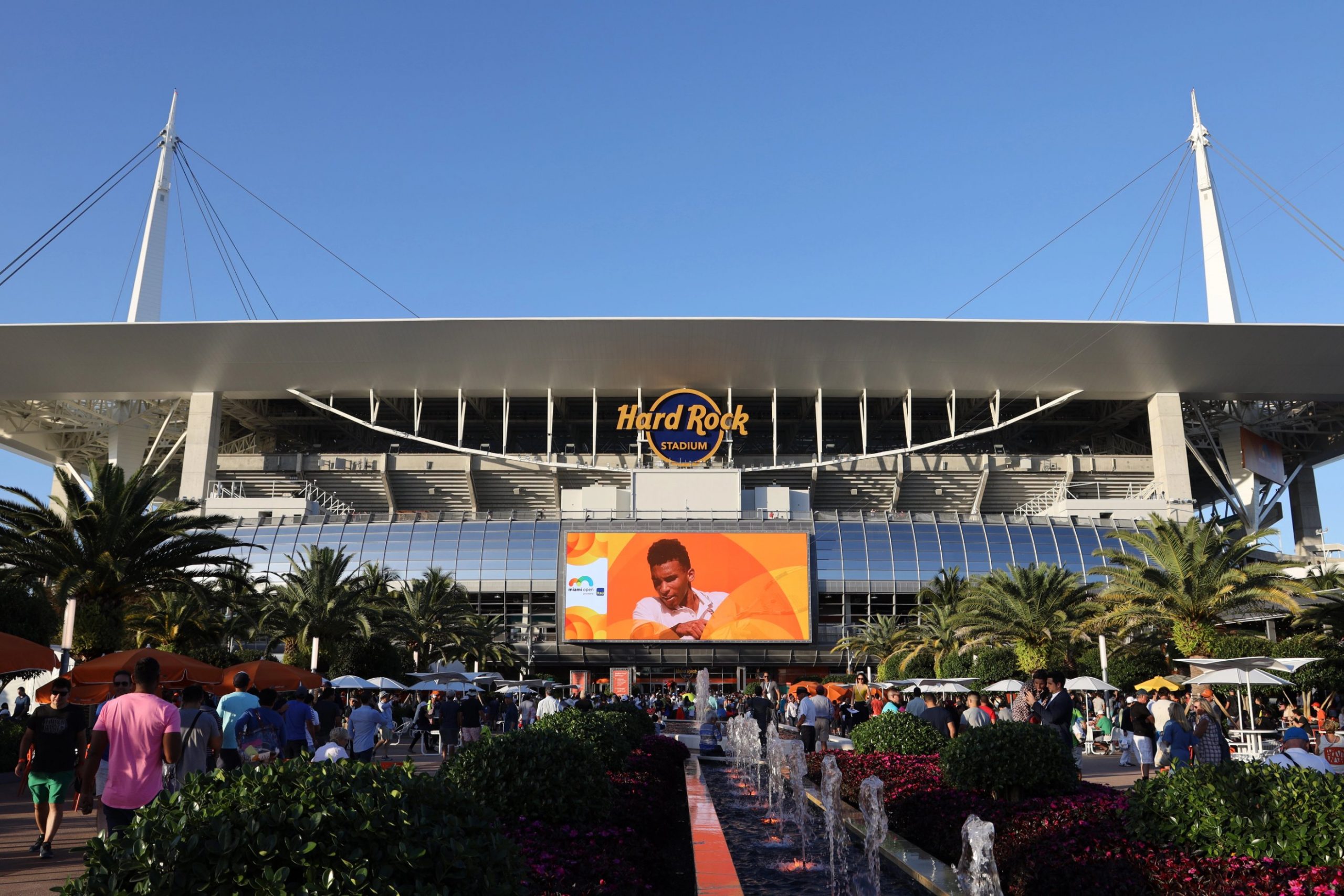 Miami Dolphins - Eine Menschenmenge versammelt sich an einem sonnigen Tag vor dem Hard Rock Stadium. Palmen, Springbrunnen und eine große Leinwand, auf der Ryan Crow in einem weißen Hemd zu sehen ist, ragen unter dem ikonischen Dach des Stadions und dem "Hard Rock Stadium"-Schild in den klaren blauen Himmel. Diese Beschreibung wurde mit der FootballR KI automatisch generiert.