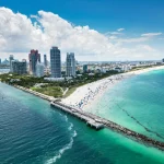 Luftaufnahme einer pulsierenden Stadtlandschaft am Strand mit hohen modernen Gebäuden, wo die Spannung der NFL Preseason entlang des weißen Sandstrandes mit vielen Sonnenanbetern und Sonnenschirmen, türkisfarbenem Meerwasser und einem steinernen Steg unter einem strahlend blauen Himmel zu spüren ist. Diese Beschreibung wurde mit der FootballR KI automatisch generiert.