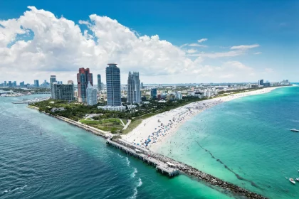 Luftaufnahme einer pulsierenden Stadtlandschaft am Strand mit hohen modernen Gebäuden, wo die Spannung der NFL Preseason entlang des weißen Sandstrandes mit vielen Sonnenanbetern und Sonnenschirmen, türkisfarbenem Meerwasser und einem steinernen Steg unter einem strahlend blauen Himmel zu spüren ist. Diese Beschreibung wurde mit der FootballR KI automatisch generiert.