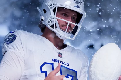 Josh Allen in einem weißen Buffalo NFL Rivalries-Trikot mit der Nummer 17 steht mit einem Helm im fallenden Schnee. Er hält einen großen Schneeball statt eines Footballs und schaut konzentriert, während Schneeflocken aufgewirbelt werden und eine winterliche Atmosphäre schaffen. Diese Beschreibung wurde mit der FootballR KI automatisch generiert.
