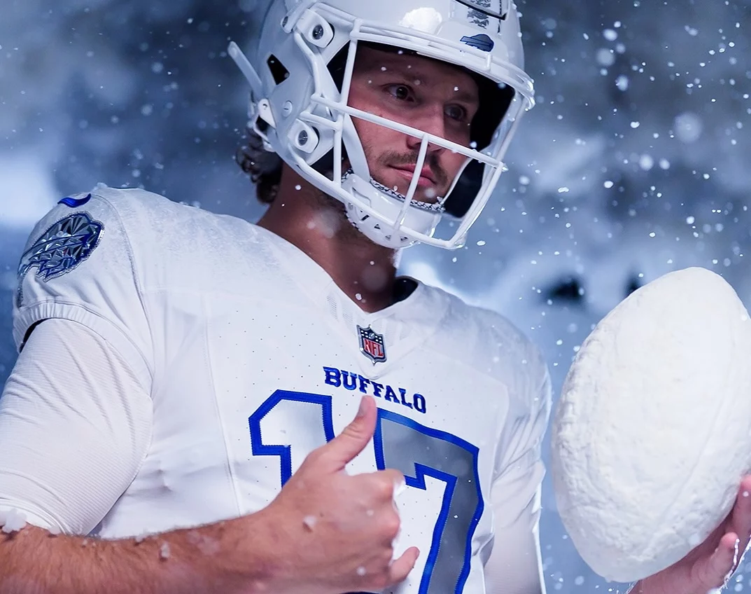 Josh Allen in einem weißen Buffalo NFL Rivalries-Trikot mit der Nummer 17 steht mit einem Helm im fallenden Schnee. Er hält einen großen Schneeball statt eines Footballs und schaut konzentriert, während Schneeflocken aufgewirbelt werden und eine winterliche Atmosphäre schaffen. Diese Beschreibung wurde mit der FootballR KI automatisch generiert.