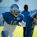 Ein Detroit Lions-Footballspieler mit silbernem Helm und blauem Trikot mit der Nummer 10 übt das Blocken gegen ein blaues gepolstertes Schild, das von einem Trainer während einer sonnigen Trainingseinheit im Freien in der NFC North gehalten wird. Eine weitere Person ist unscharf im Hintergrund zu erkennen. Diese Beschreibung wurde mit der FootballR KI automatisch generiert.