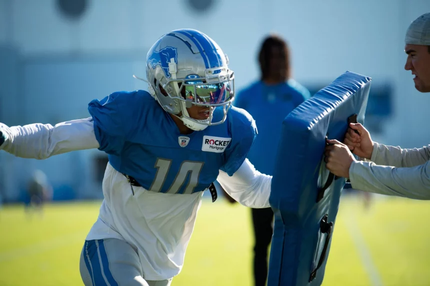 Ein Detroit Lions-Footballspieler mit silbernem Helm und blauem Trikot mit der Nummer 10 übt das Blocken gegen ein blaues gepolstertes Schild, das von einem Trainer während einer sonnigen Trainingseinheit im Freien in der NFC North gehalten wird. Eine weitere Person ist unscharf im Hintergrund zu erkennen. Diese Beschreibung wurde mit der FootballR KI automatisch generiert.