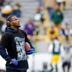 Ein Footballspieler, der Micah Parsons ähnelt, trägt Kopfhörer, ein schwarzes "Throw to Diggs"-T-Shirt über einem blauen Kapuzenpulli und blaue Handschuhe, während er auf dem Feld steht und einen Football hält. Unscharfe Stadionsitze und Fans in grünen Trikots füllen den Hintergrund. Diese Beschreibung wurde mit der FootballR KI automatisch generiert.