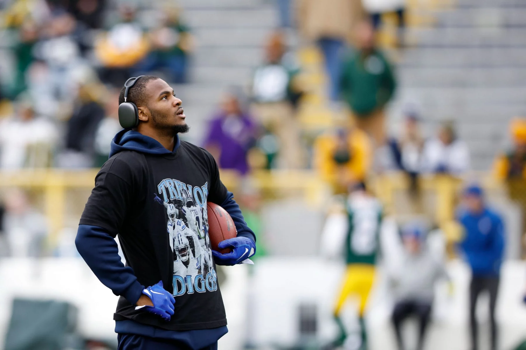 Ein Footballspieler, der Micah Parsons ähnelt, trägt Kopfhörer, ein schwarzes "Throw to Diggs"-T-Shirt über einem blauen Kapuzenpulli und blaue Handschuhe, während er auf dem Feld steht und einen Football hält. Unscharfe Stadionsitze und Fans in grünen Trikots füllen den Hintergrund. Diese Beschreibung wurde mit der FootballR KI automatisch generiert.
