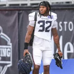 Ein Football-Spieler, Lonnie Johnson Jr., der eine Trainingsuniform der Houston Texans mit der Nummer 32 trägt, steht im Freien und hält einen Helm und Stollenschuhe. Er hat Dreadlocks, sichtbare Tattoos und Schulterpolster. Im Hintergrund ist ein großes schwarzes "Training Camp"-Banner zu sehen. Diese Beschreibung wurde mit der FootballR KI automatisch generiert.