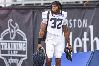 Ein Football-Spieler, Lonnie Johnson Jr., der eine Trainingsuniform der Houston Texans mit der Nummer 32 trägt, steht im Freien und hält einen Helm und Stollenschuhe. Er hat Dreadlocks, sichtbare Tattoos und Schulterpolster. Im Hintergrund ist ein großes schwarzes "Training Camp"-Banner zu sehen. Diese Beschreibung wurde mit der FootballR KI automatisch generiert.