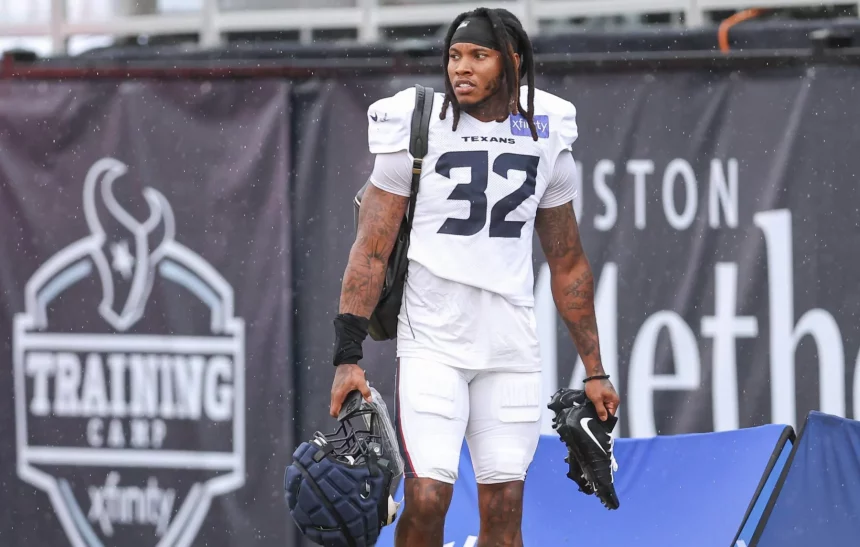 Ein Football-Spieler, Lonnie Johnson Jr., der eine Trainingsuniform der Houston Texans mit der Nummer 32 trägt, steht im Freien und hält einen Helm und Stollenschuhe. Er hat Dreadlocks, sichtbare Tattoos und Schulterpolster. Im Hintergrund ist ein großes schwarzes "Training Camp"-Banner zu sehen. Diese Beschreibung wurde mit der FootballR KI automatisch generiert.
