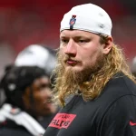 Ein ernster Kaleb McGary mit langen blonden Haaren und Bart trägt eine weiße Mütze und ein schwarzes Atlanta-Falcons-Shirt. Er steht in einem Innenraum, während im Hintergrund unscharfe Mannschaftskameraden in schwarzer Kleidung zu sehen sind. Diese Beschreibung wurde mit der FootballR KI automatisch generiert.