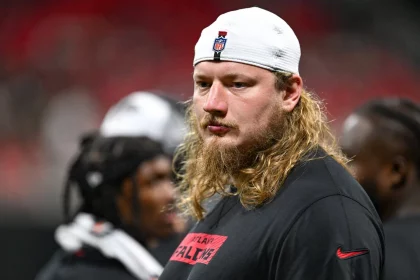 Ein ernster Kaleb McGary mit langen blonden Haaren und Bart trägt eine weiße Mütze und ein schwarzes Atlanta-Falcons-Shirt. Er steht in einem Innenraum, während im Hintergrund unscharfe Mannschaftskameraden in schwarzer Kleidung zu sehen sind. Diese Beschreibung wurde mit der FootballR KI automatisch generiert.